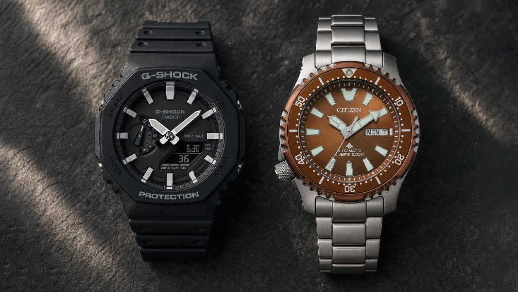 GA-2100 and Citizen Promaster side by side, split flat-lay, rugged dark surface 